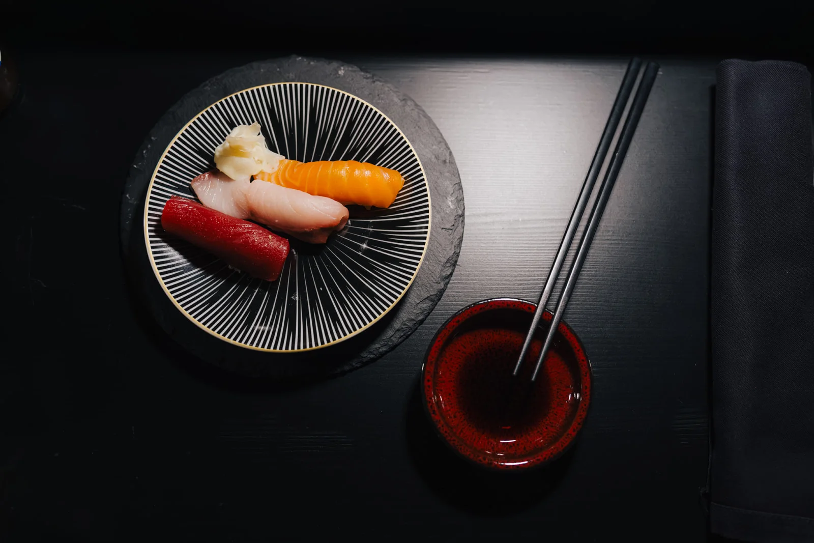 Nigiri auf gestreiftem Teller über Schiefer, daneben die rote Schale Sojasauce mit Stäbchen auf schwarzem Tisch