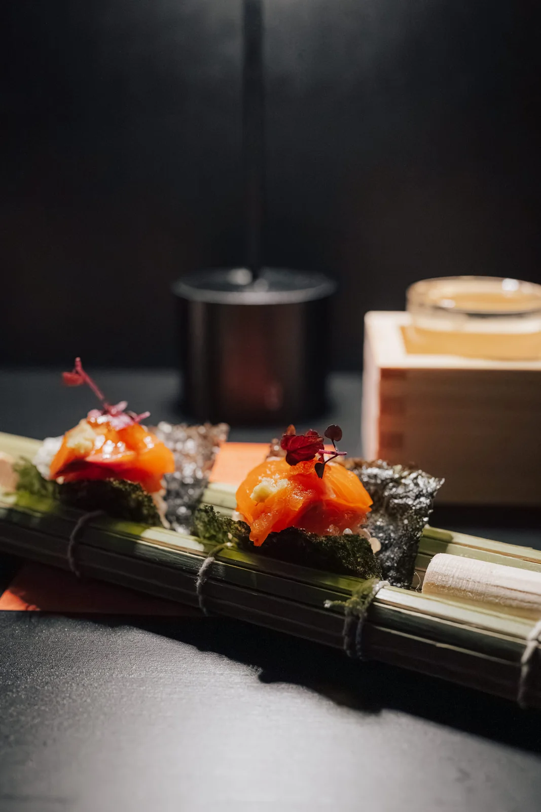 Lachs auf Nori mit Blüten, auf der Bambusmatte angerichtet, dahinter ein Sakebecher